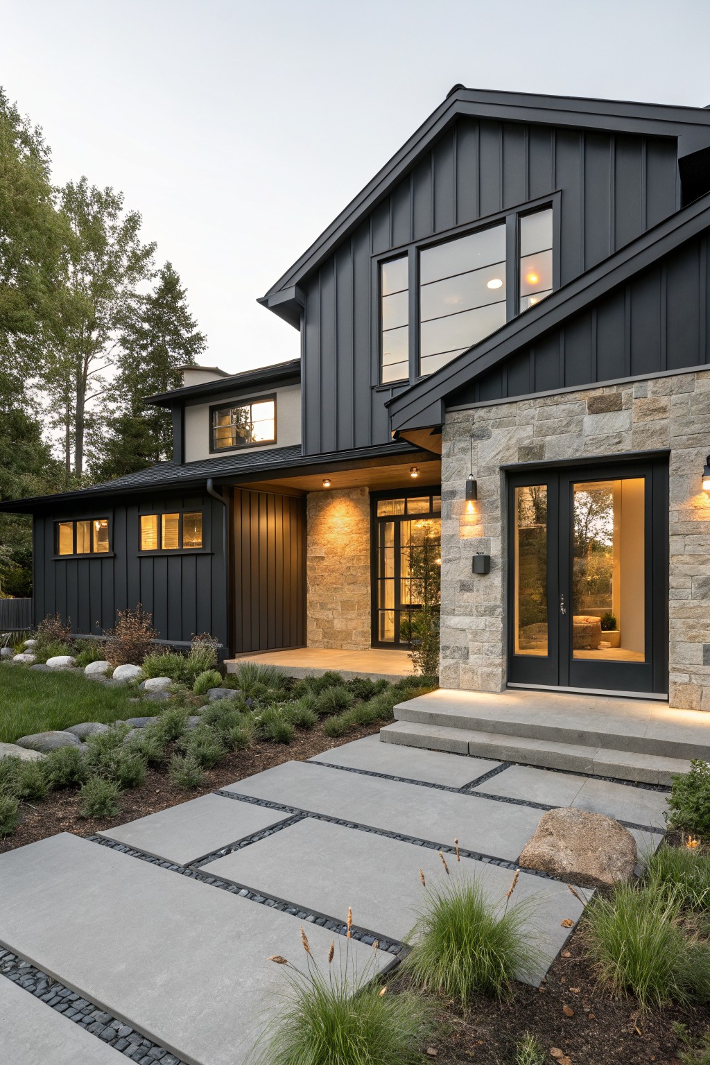 Modern farmhouse house exterior at dusk with black board-and-batten siding, stone masonry around black double doors, wood accents, concrete steps and path, low plants, rocks, and trees in yard.