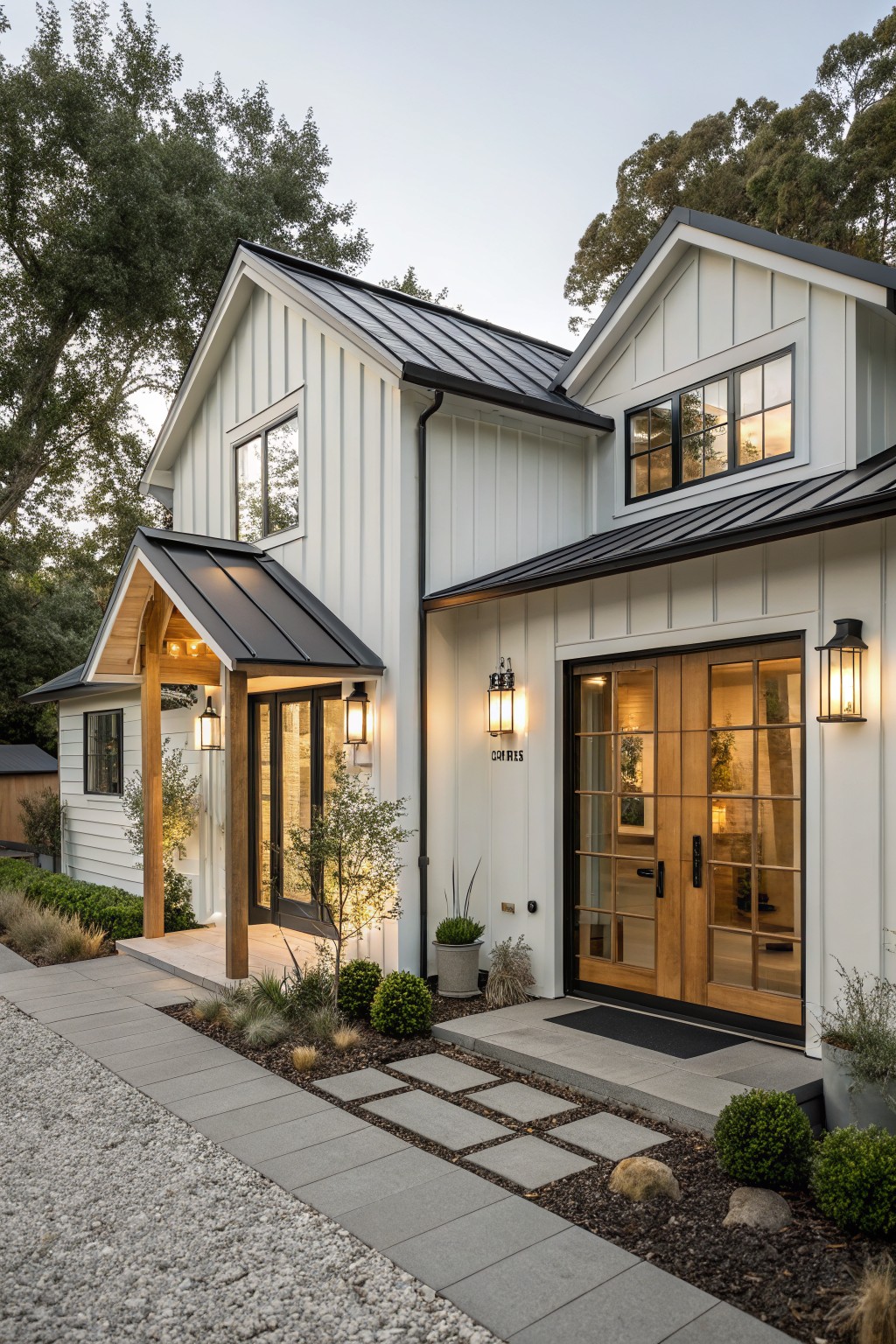 Two-story modern farmhouse exterior with white board-and-batten siding, black metal roof sections, wooden double doors, lanterns, and gravel driveway edged by low plants and stepping stones at dusk.