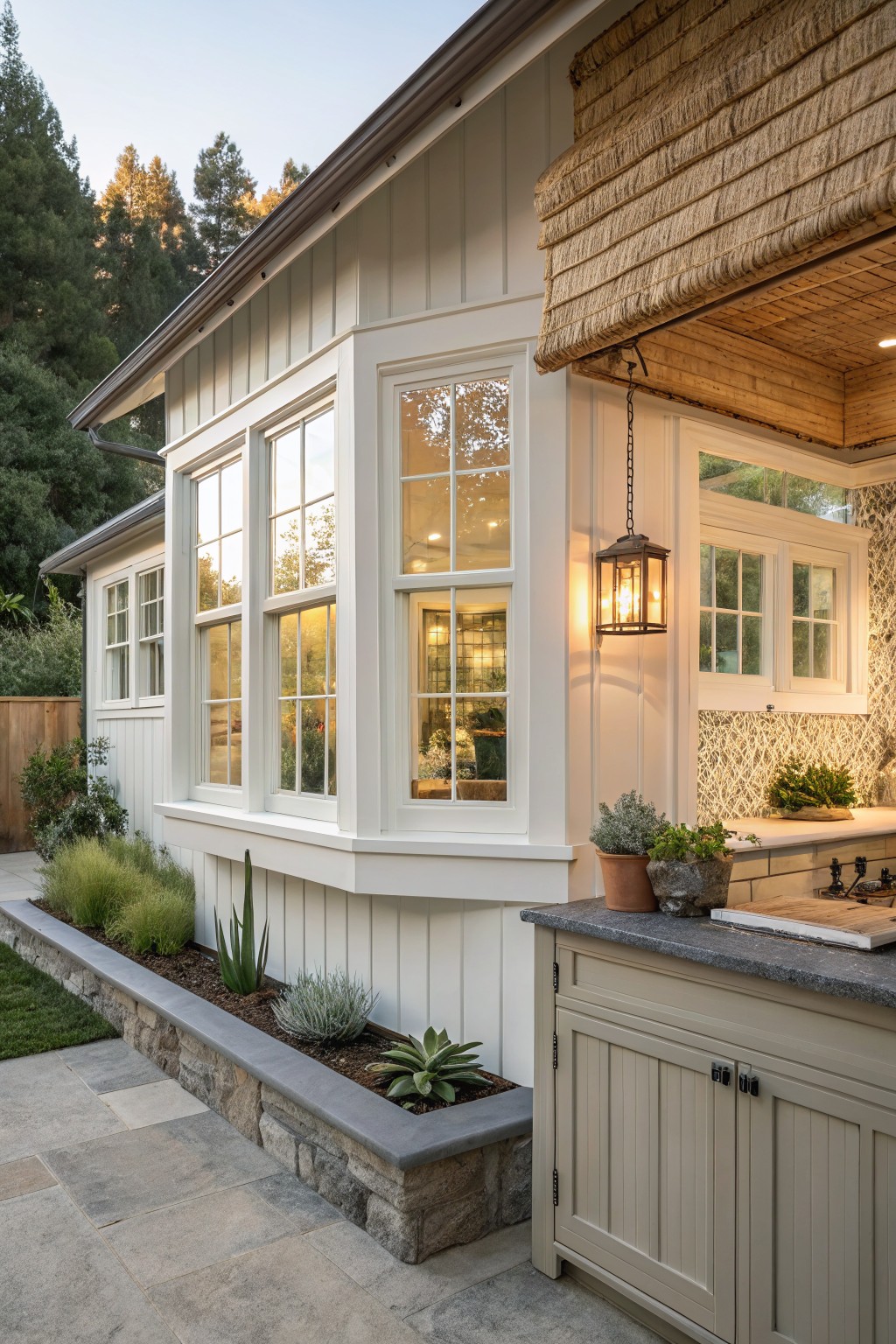 White board-and-batten house exterior featuring a protruding bay window with multiple panes next to an outdoor kitchen counter with cabinets, a hanging lantern light, and a stone-edged planting bed with succulents.