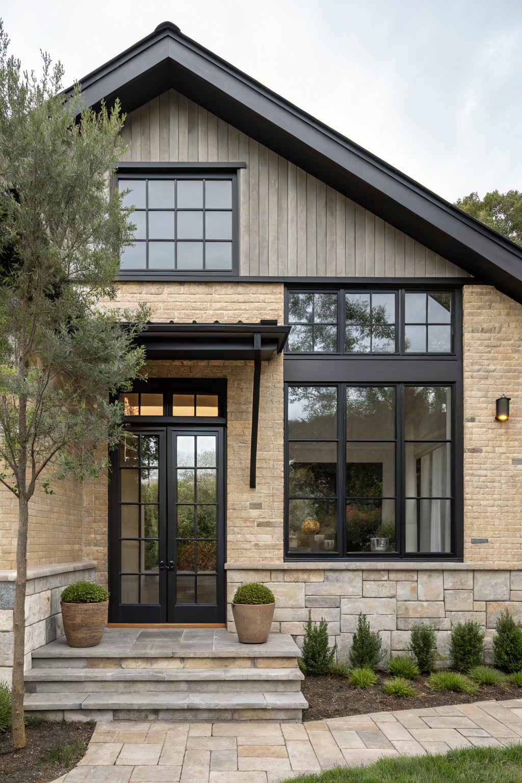 Front exterior of a two-story modern farmhouse house with beige brick lower walls, vertical cedar siding on the upper gable, black metal-framed grid windows, double glass entry doors under a black awning, stone steps, potted plants, and an olive tree.