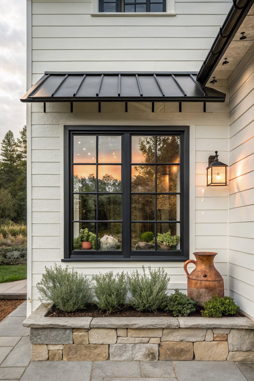 White clapboard house exterior with black standing seam metal awning over a large black-framed grid window, illuminated lantern light beside it, potted plants inside the window, large terracotta pot, and stone planter box filled with shrubs on a paved path.