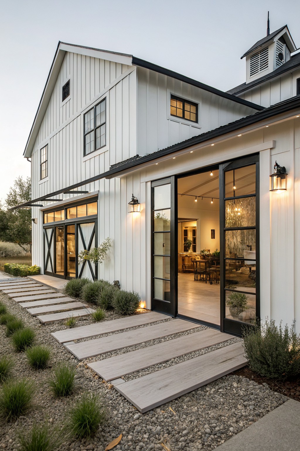 White board and batten farmhouse exterior with large open black sliding doors revealing interior dining area, flanked by black framed windows, stone pathway leading to entry, and low landscaping along gravel ground.