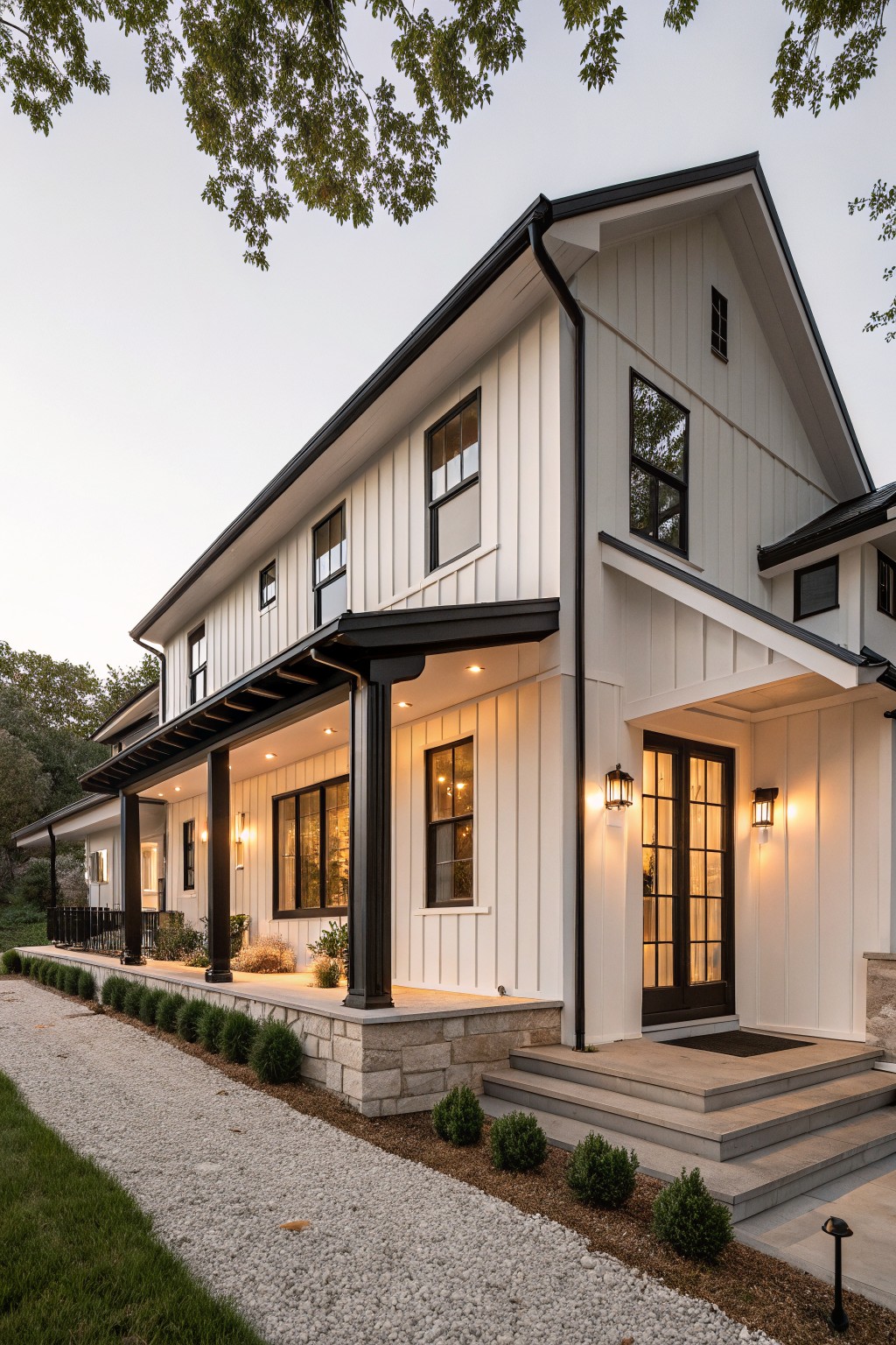 Side exterior view of a two-story modern farmhouse featuring white board-and-batten siding, black window and door trim, covered porch with black posts, stone foundation accents, gravel pathway, and low shrubs.