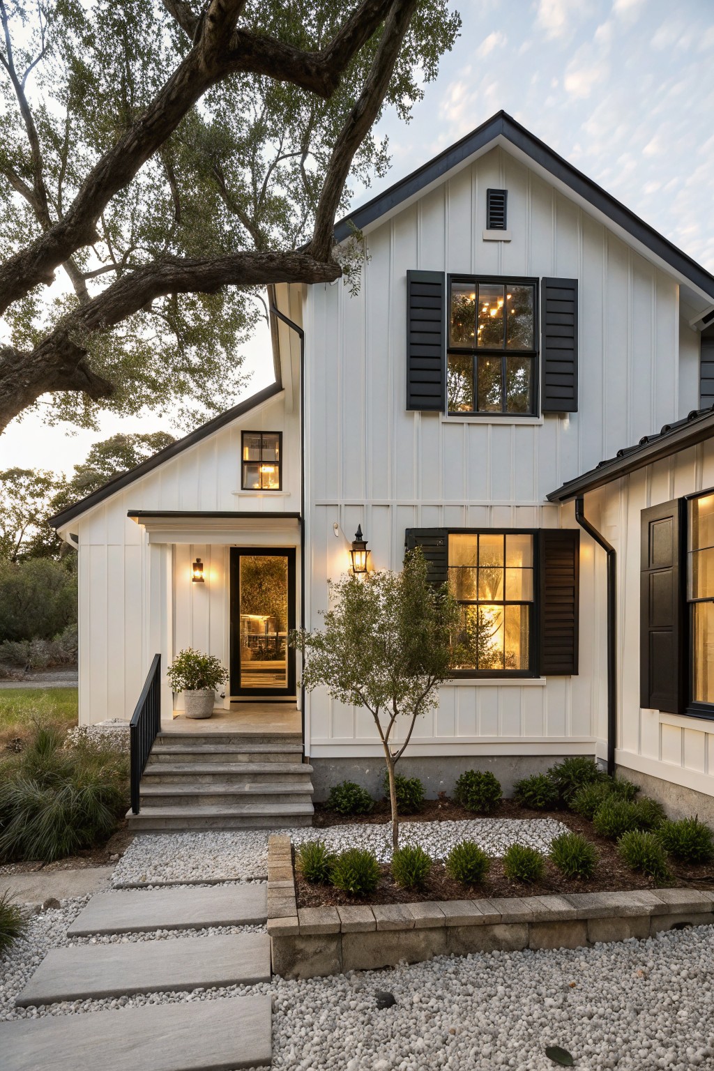 White two-story modern farmhouse exterior with black shutters, trim, roof, and lanterns on the front porch, large overhanging oak tree, gravel landscaping, and stone steps.