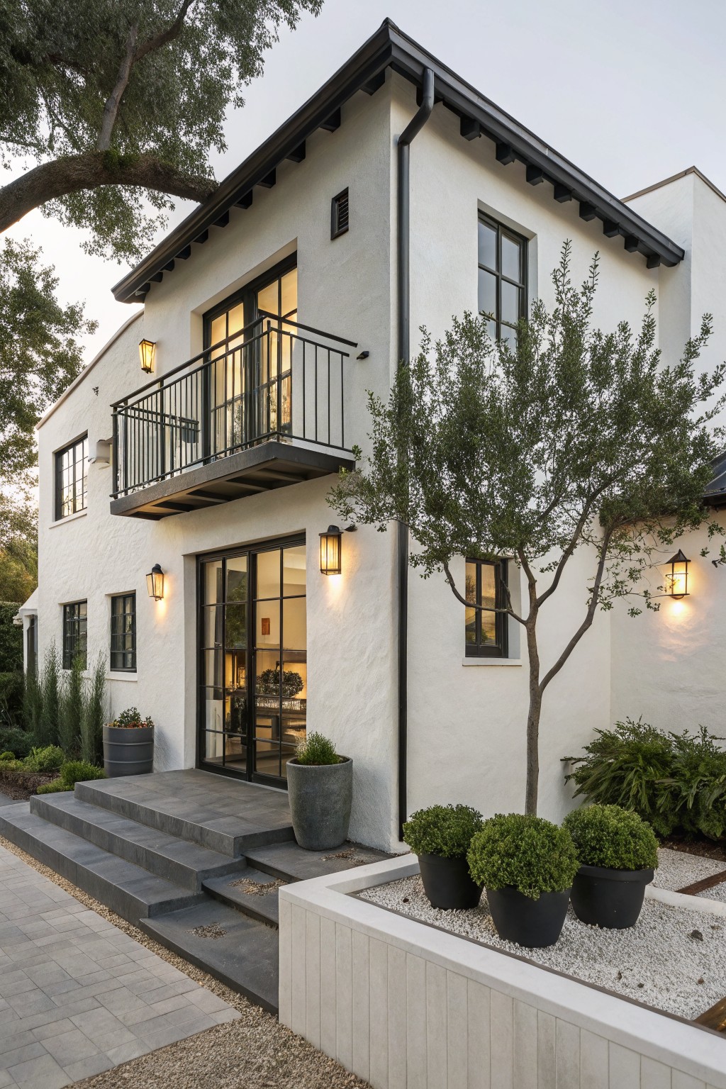 Corner view of a two-story white stucco house featuring black-framed windows, doors, balcony railing, and trim, with stone steps leading to a glass entry door flanked by lanterns and potted plants on a paved patio.