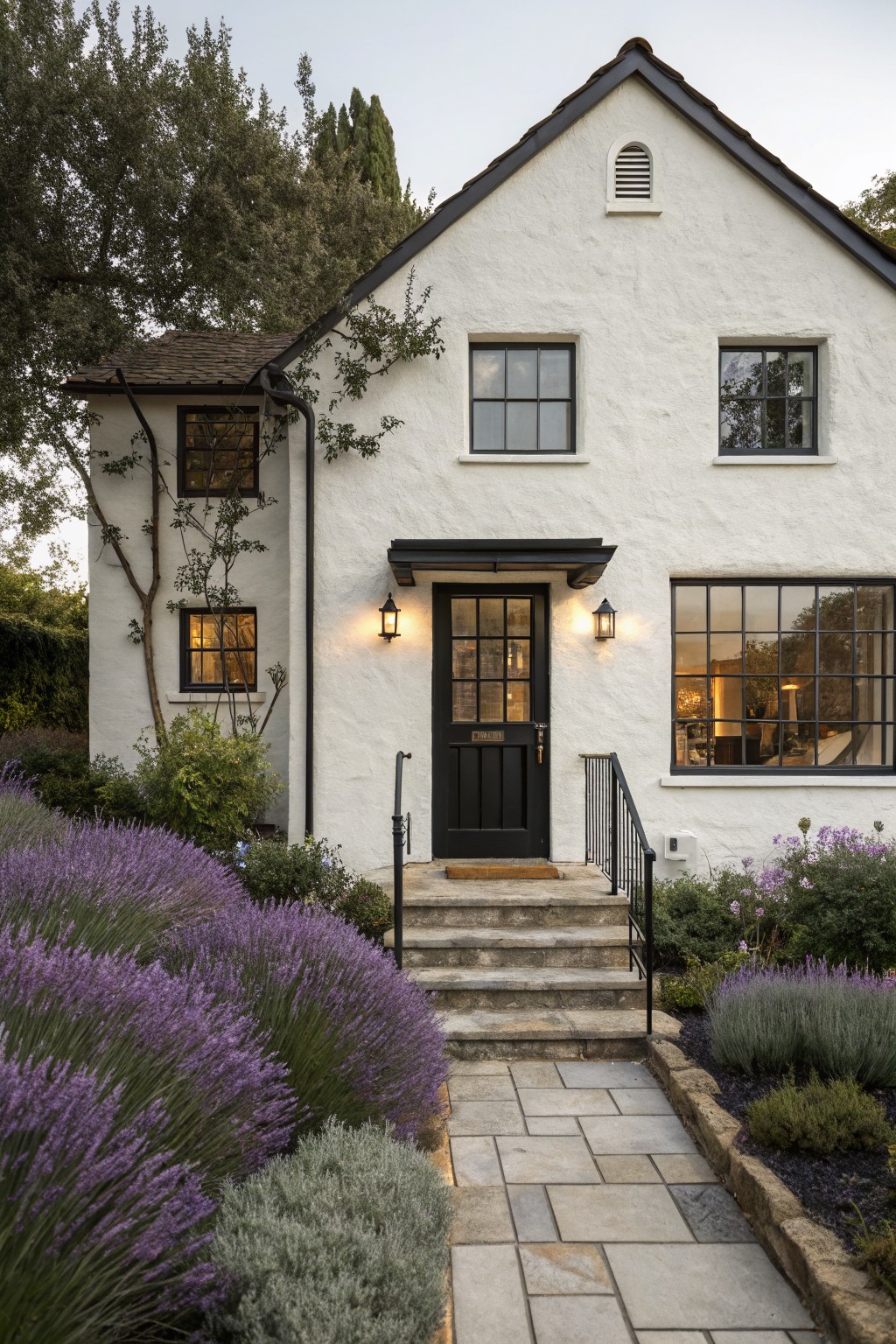 White stucco house exterior with black-framed windows, black front door, lantern lights, stone steps, and purple lavender plants bordering a flagstone path.