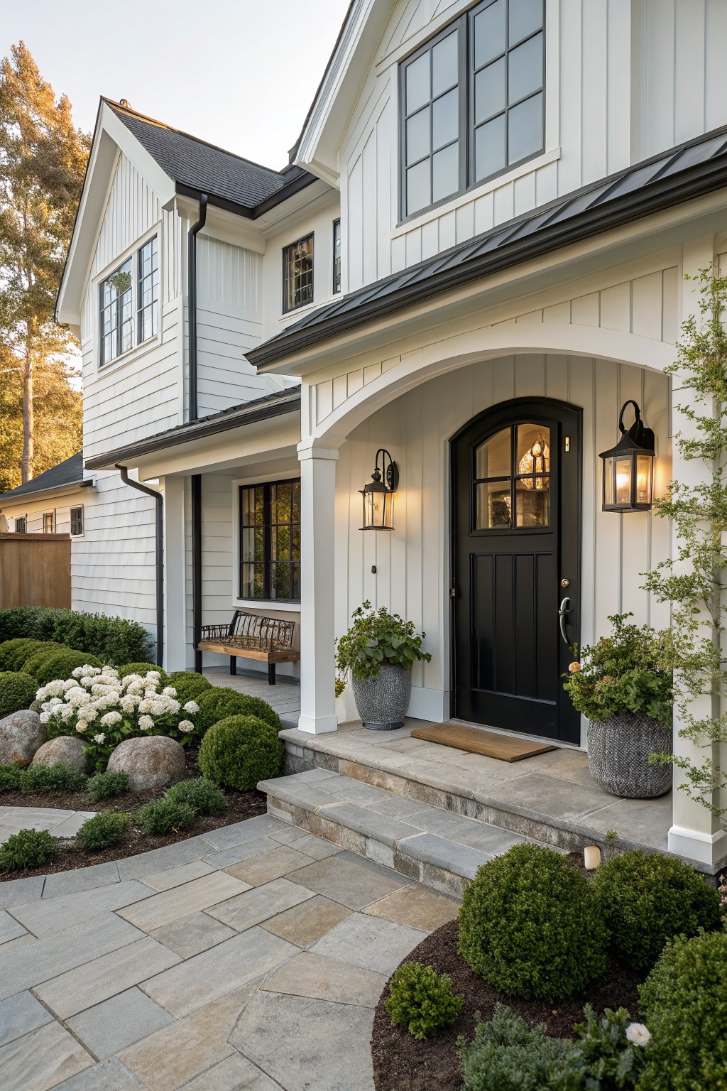 White shiplap modern farmhouse with black arched front door under porch, flanked by lanterns and bench, curved bluestone path edged by boulders, hydrangeas, boxwoods, and potted plants leading to steps.