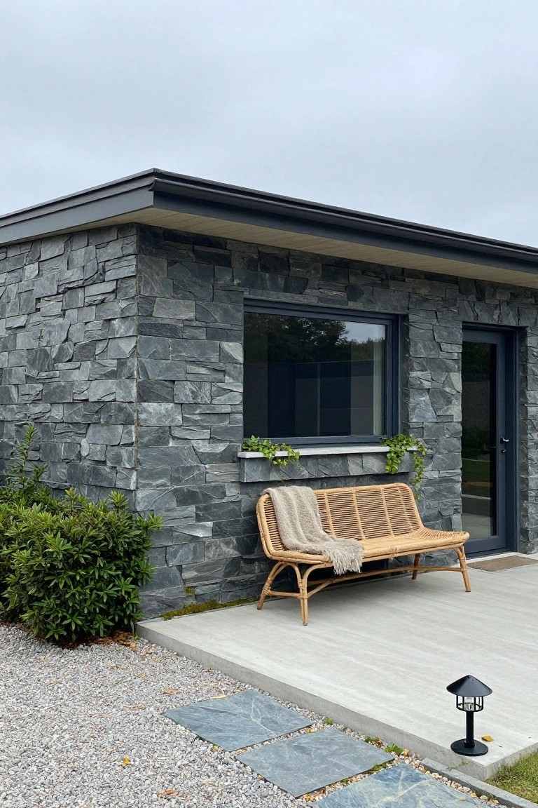 Corner view of a modern house with dark slate stone walls, black-framed window and glass door, rattan bench with gray blanket on a concrete platform, gravel ground, low shrubs, and a black lantern nearby.