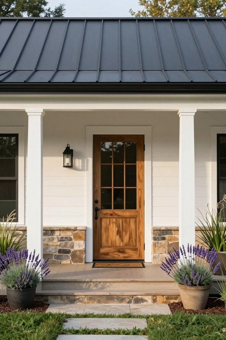 Front view of a white board-and-batten sided house with dark standing-seam metal roof, covered porch supported by white columns on stone bases, wooden door with nine glass panes, wall lantern, and potted lavender plants flanking stone steps leading to a wood porch floor.