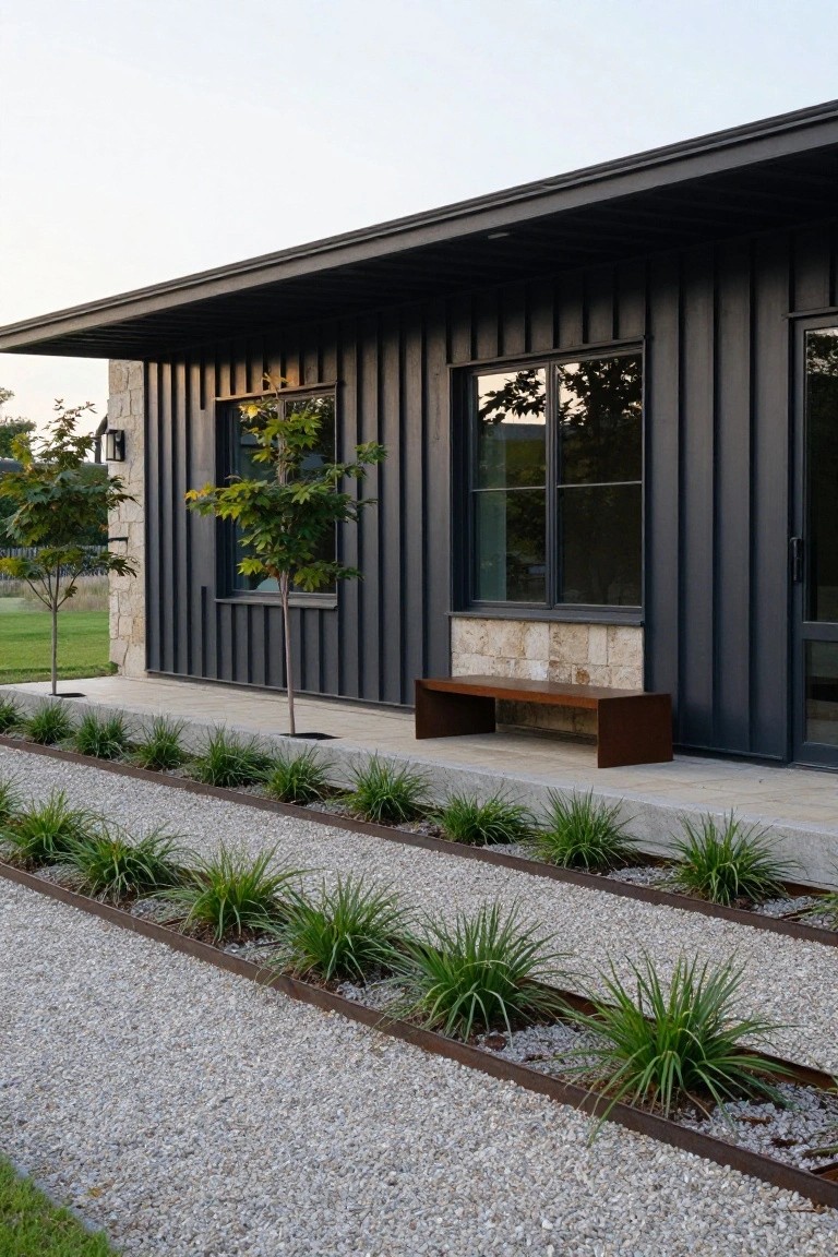 Side view of a modern house with black corrugated metal siding, large windows, stone base, wooden bench, and gravel beds edged with metal strips planted with grasses.
