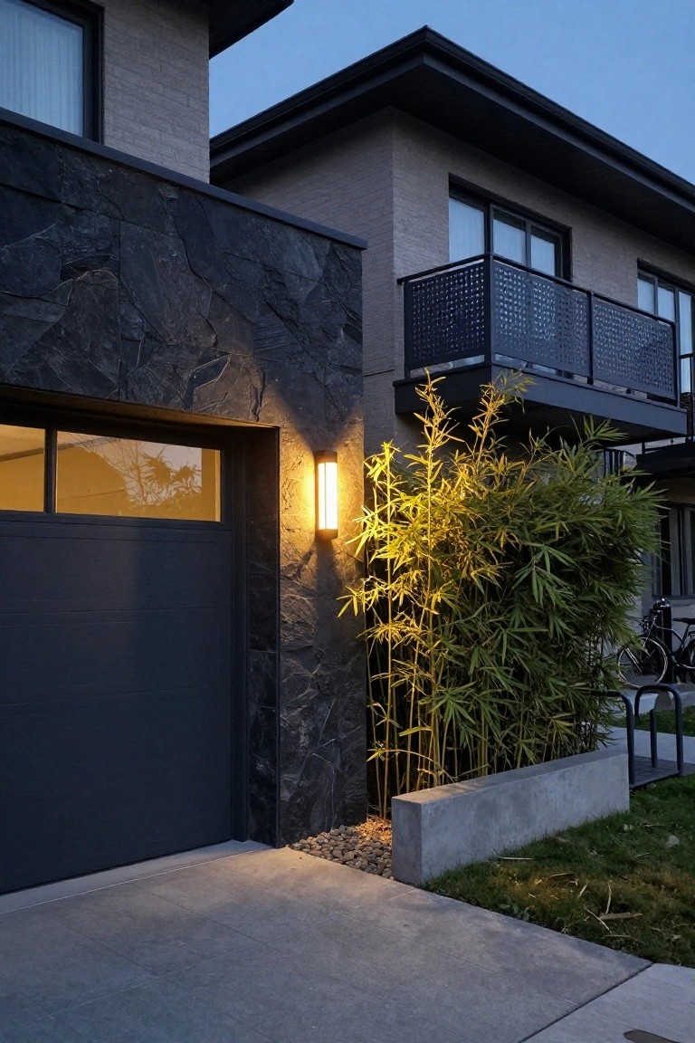 Modern house exterior at dusk showing a dark stone veneer wall next to a dark gray garage door with glass panels, a tall yellow wall light, green bamboo plants in a concrete-edged bed, and bike racks on a concrete driveway.