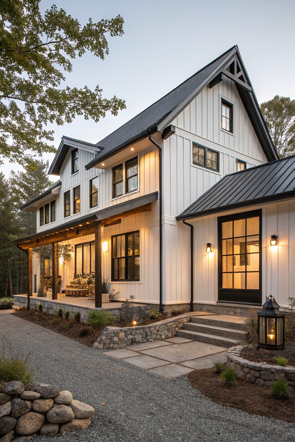 Two-story house exterior with white vertical board-and-batten siding, black-framed windows and doors, dark metal gable roof, wraparound porch, stone steps, gravel driveway, and low landscaping at dusk.