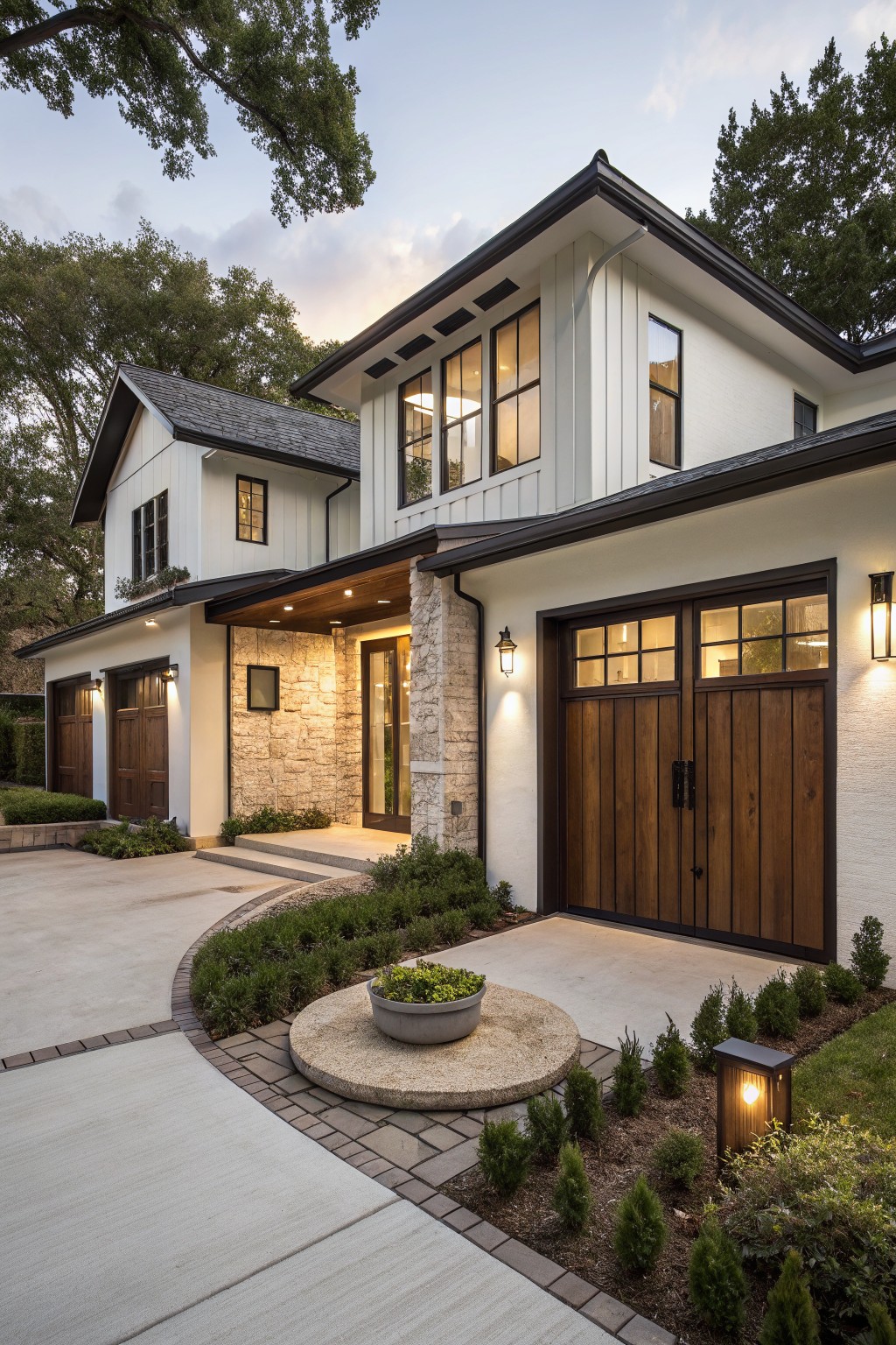 White board-and-batten sided modern farmhouse exterior with stone entry, wood garage doors, curved concrete driveway, low shrubs, and path lighting at dusk.