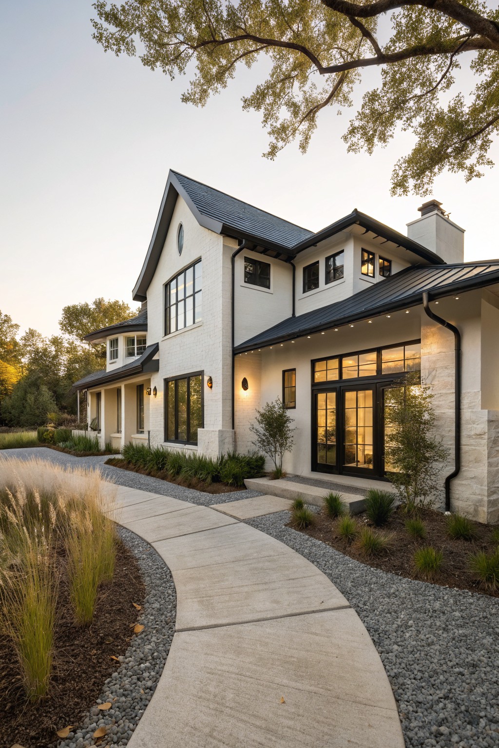 Side exterior view of a two-story white brick house with black metal roof and trim, large windows, glass entry doors with stone surround, curved concrete pathway edged by ornamental grasses and pebbles, and trees nearby at dusk.