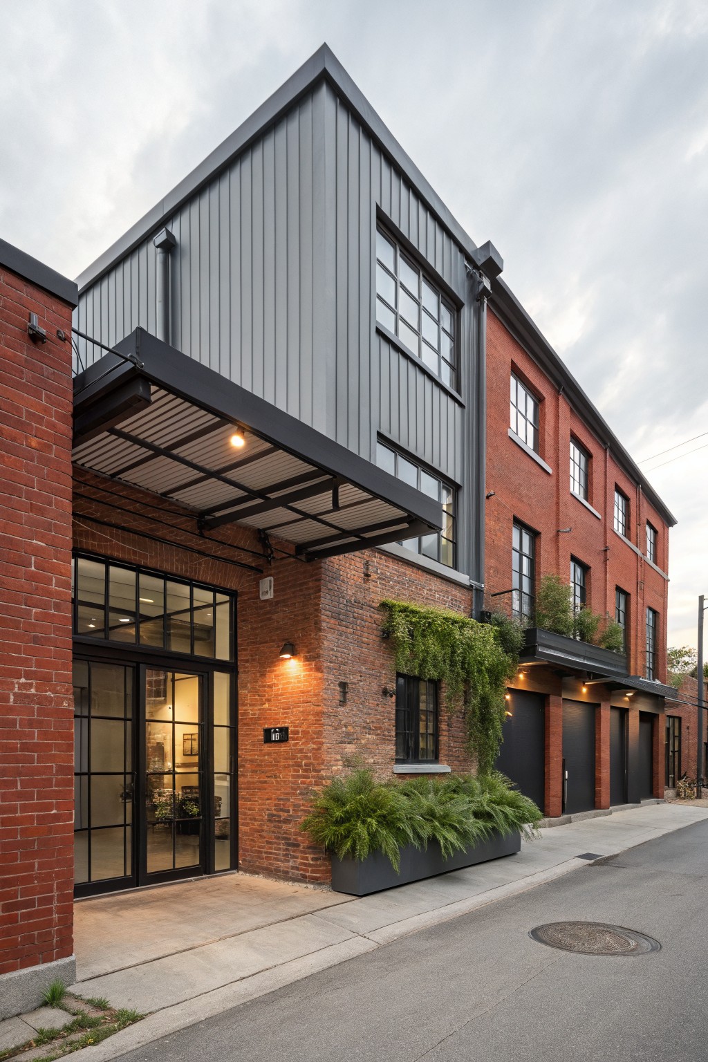 A two-story house exterior with red brick on the ground level walls and entry area, gray corrugated metal siding on the upper stories and addition, black metal canopy over a glass door entrance, black garage doors, potted ferns along the front, and a concrete sidewalk on a street.