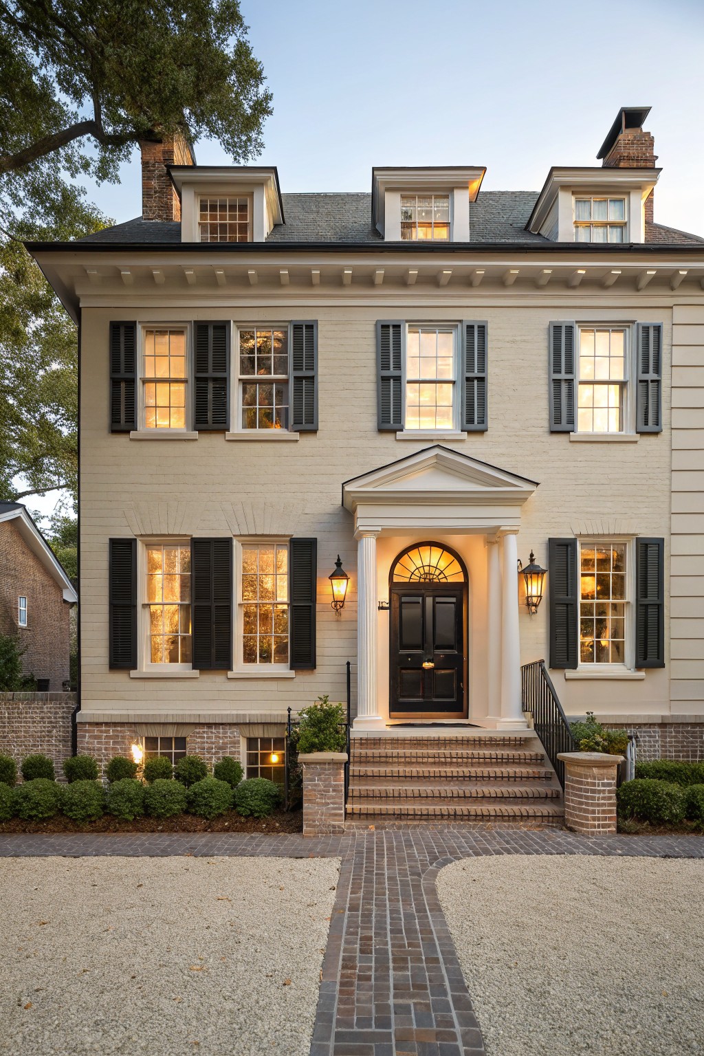 Two-story beige painted brick house with black shutters on multipane windows, white columned portico sheltering a black front door, brick steps, boxwood shrubs, and a brick path curving to a gravel driveway at evening light.