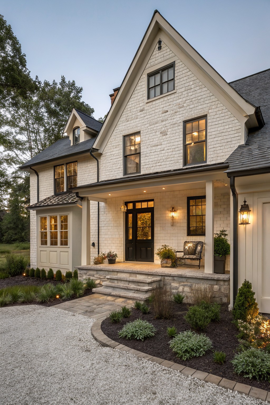 Two-story house exterior featuring white painted brick on main walls, cream vertical siding on garage, black-framed windows and front door, covered porch with columns, stone entry steps, gravel driveway, and low plantings around the base.