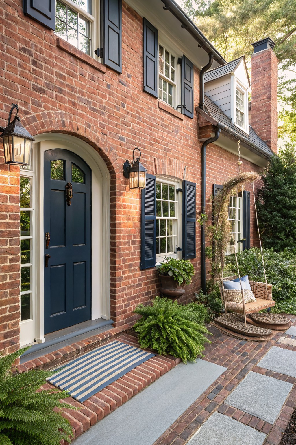 Red brick house facade featuring a navy blue arched double front door with brass hardware, matching navy shutters on white-framed windows, wall lanterns, potted plants, a hanging porch swing, ferns, and a brick pathway with concrete pavers.