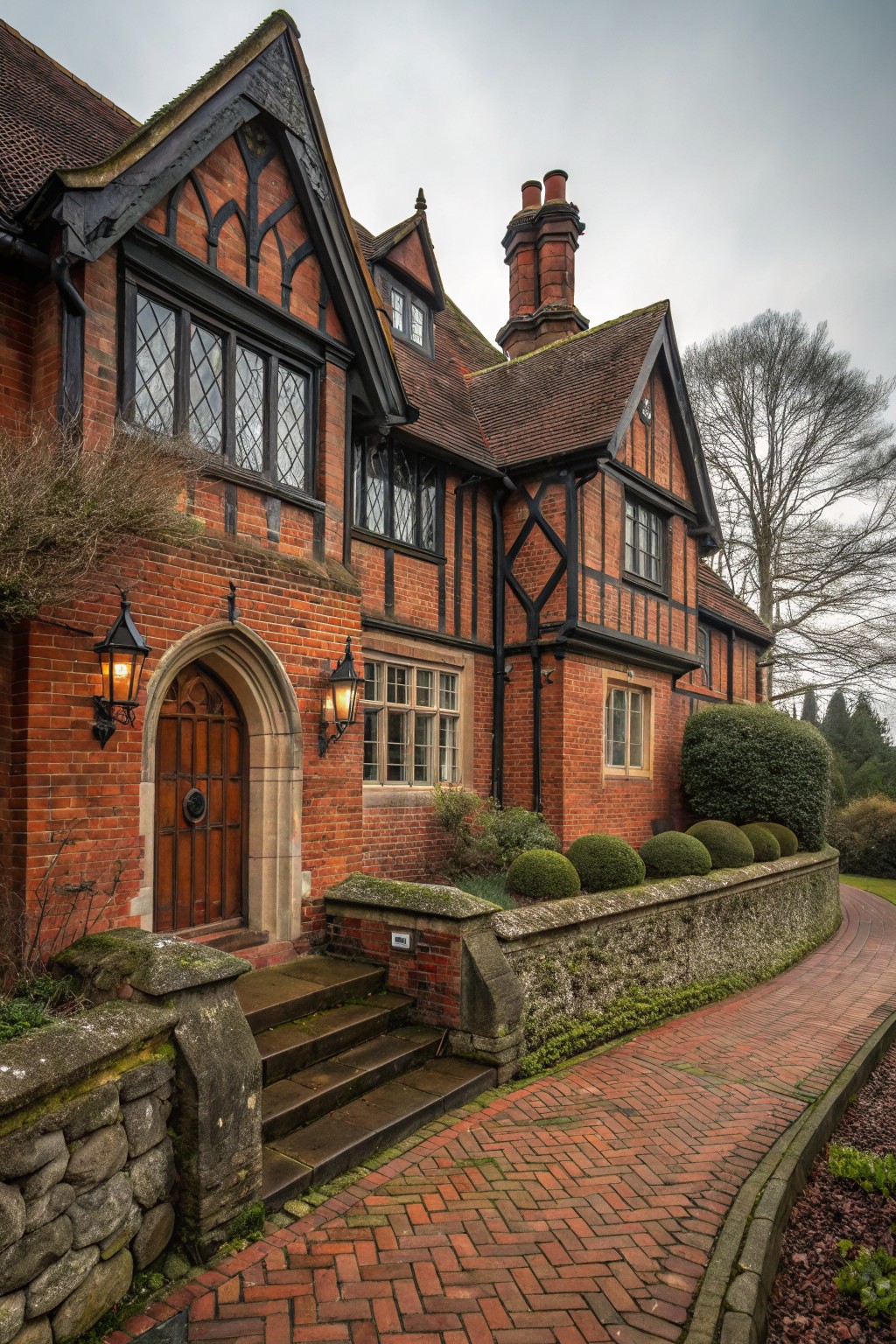 Red brick Tudor-style house exterior featuring black half-timber framing, arched wooden front door with lanterns, leaded windows, stone entry steps, clipped shrubs, and a curving red brick pathway beside a low stone wall under gray skies.