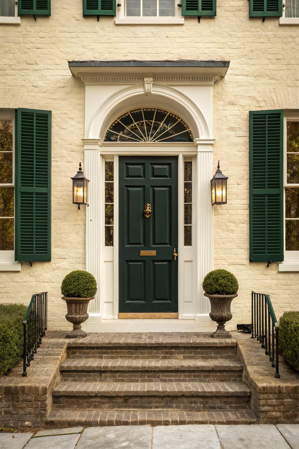 Cream painted brick house exterior with dark green front door, matching green shutters, white trim around a fanlight window, lanterns on pillars, topiary urns, and brick steps leading to the entry.