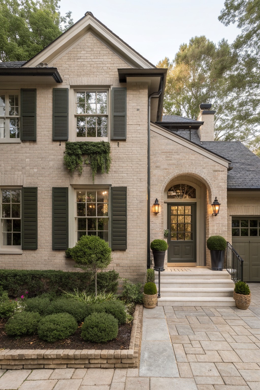 Two-story light beige brick house exterior featuring dark green shutters, an arched green front door with lanterns, boxwood shrubs in pots, and a paved entry path bordered by low hedges.