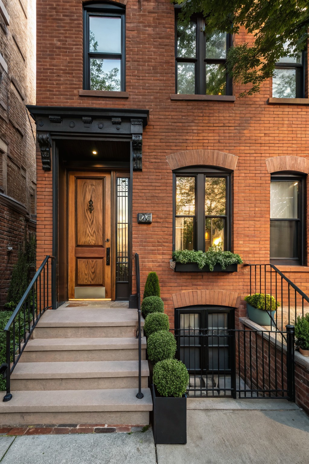 Red brick rowhouse exterior with black metal window frames, arched windows, wooden front door with anchor carving numbered 249, stone stoop flanked by black railings and potted boxwood shrubs.