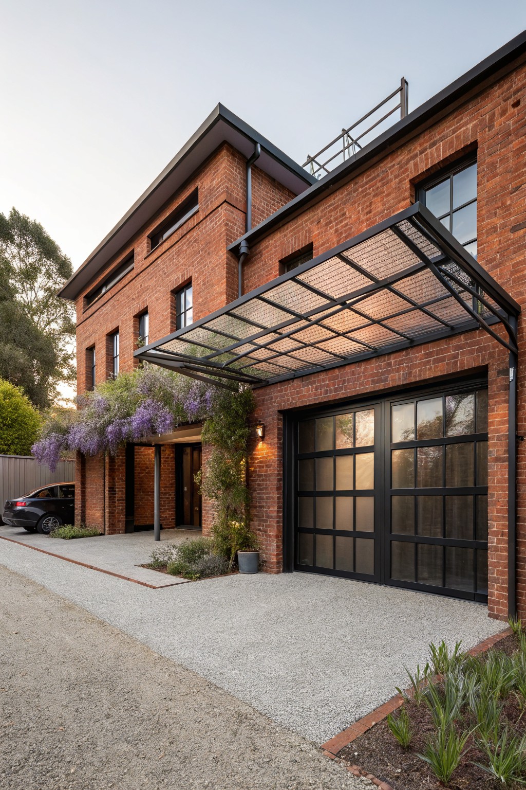 Red brick two-story house exterior featuring a large black-framed frosted glass garage door, metal-framed glass-roofed canopy extending over the driveway with purple wisteria vines, entry door, and gravel driveway.