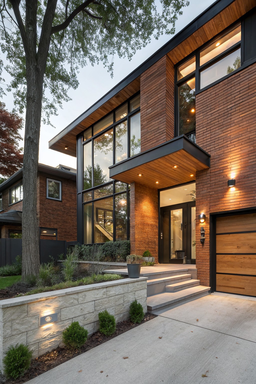 Modern two-story red brick house exterior with large glass windows, wooden cantilevered entry canopy, black metal accents, wooden garage door, stone retaining wall, and landscaping including shrubs and trees.