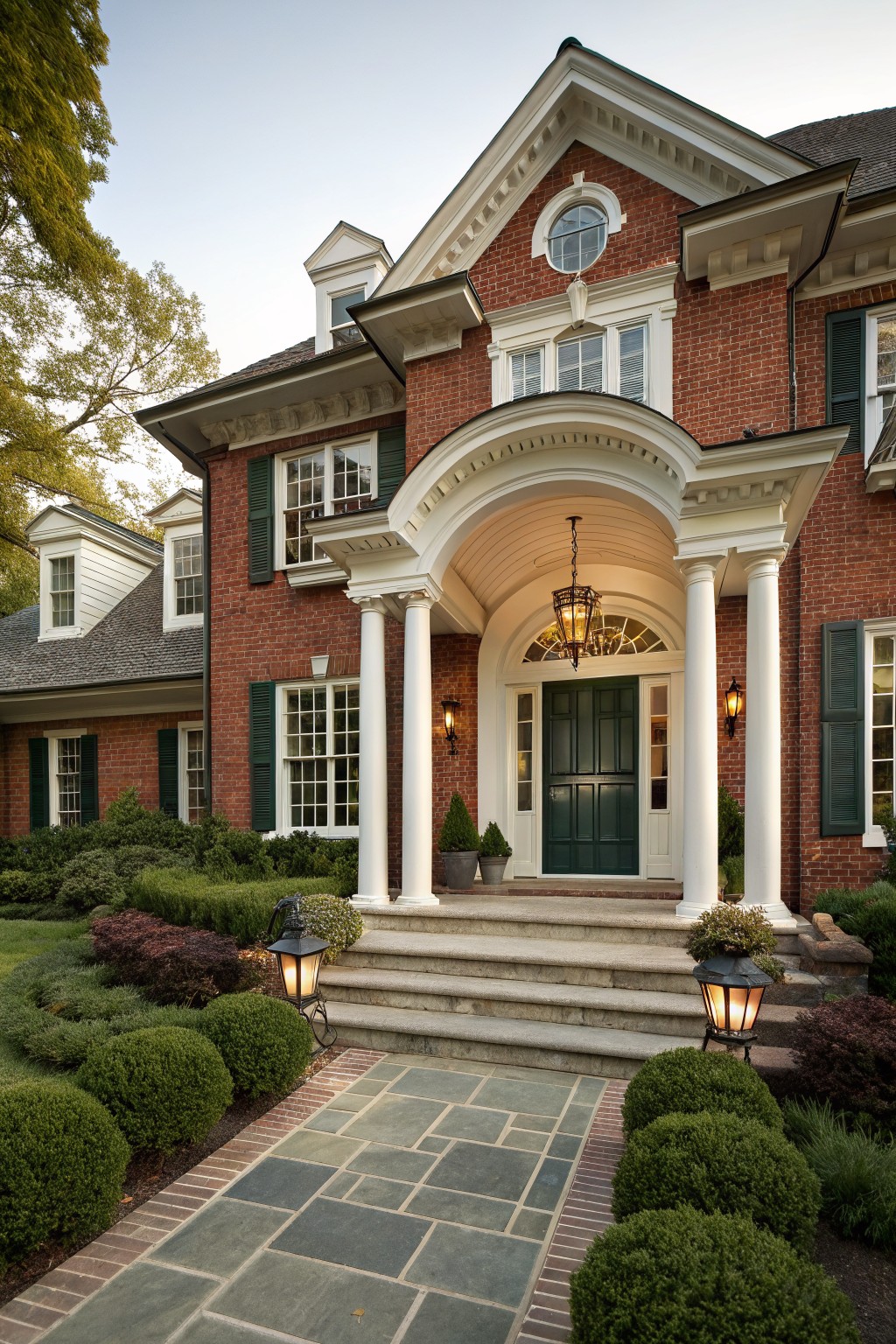 Red brick house exterior featuring a white columned portico with arched entry, green front door, lanterns, stone steps and pathway, boxwood shrubs, and trees in the yard.
