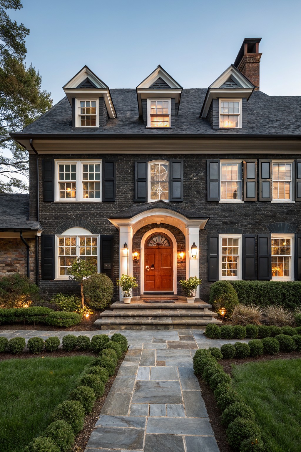 A two-story black brick Colonial Revival house with white trim, black shutters, dormer windows, a pedimented portico with red wood door, slate stone steps and pathway lined with boxwood shrubs, surrounded by lawn and trees at dusk.
