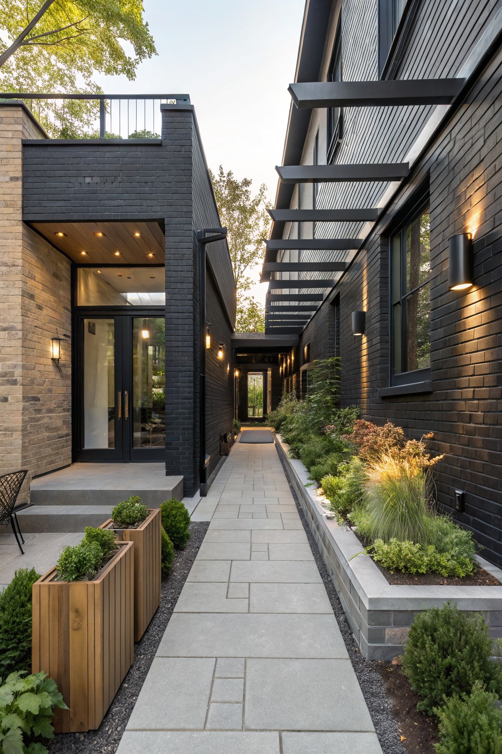 Exterior pathway of a modern black brick house with steel pergola cover, concrete pavers, wooden planters, and low greenery beds along one side leading from a glass entry door to the main structure.