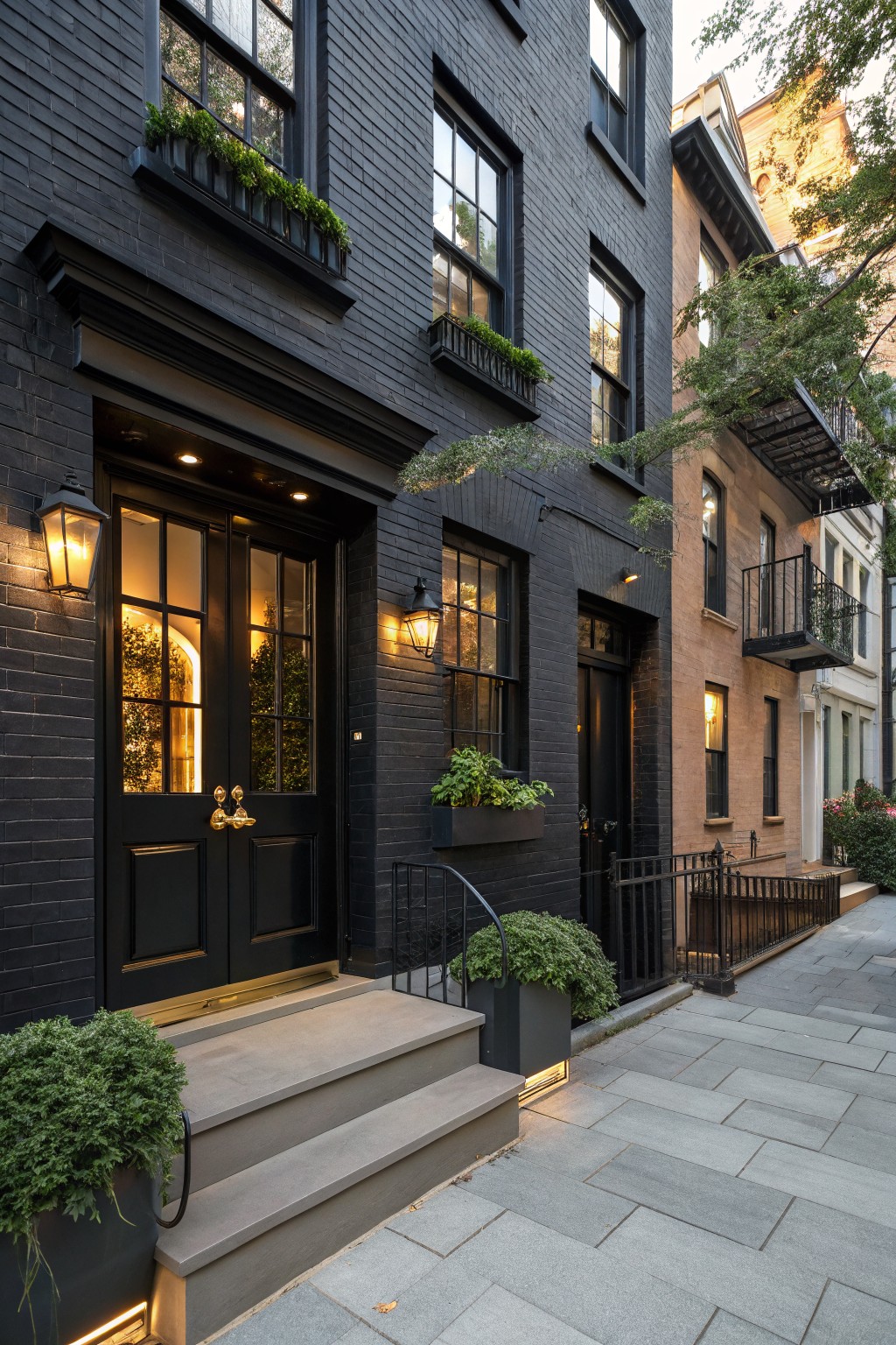 Black painted brick townhouse exterior with double black doors featuring glass panels and brass knocker, flanked by lanterns, window boxes with plants, stone steps, potted greenery, and metal railing on a city sidewalk.