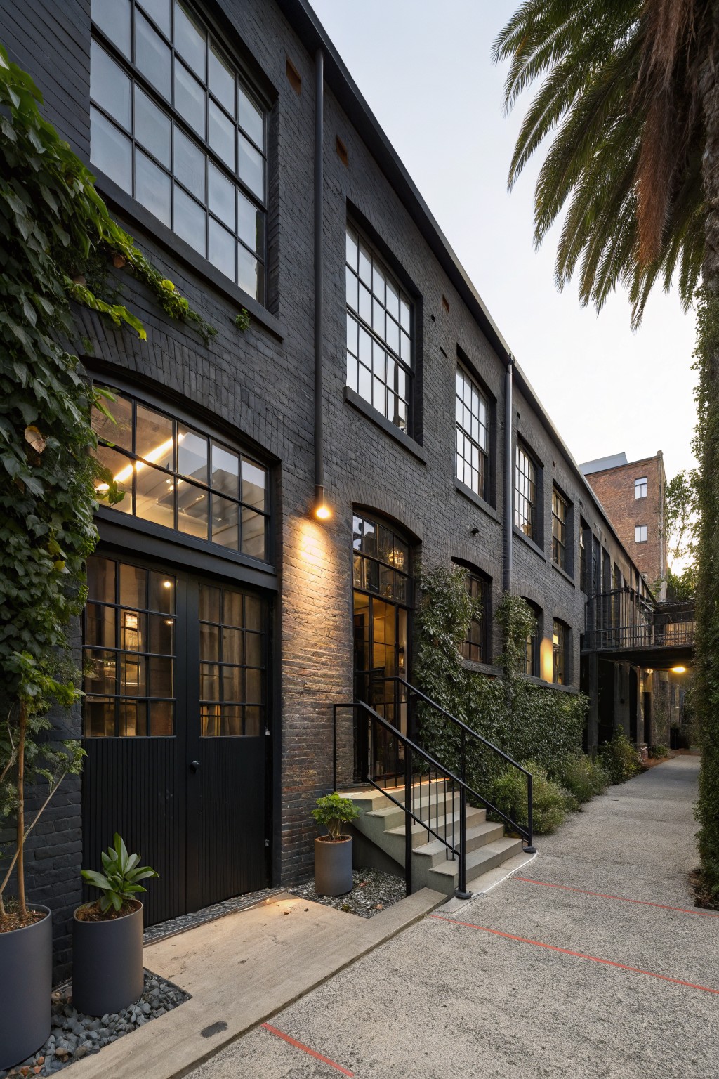 Exterior of a multi-story black brick building with large steel-framed windows, ivy climbing the walls, potted plants by black doors, metal stairs to a glass entry door, and palm trees nearby on a concrete path.