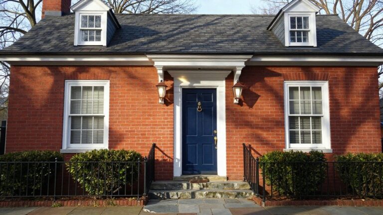 Red brick colonial-style house facade with white portico supported by columns, navy blue double front door, brass lantern lights, stone steps, and boxwood hedges on either side.