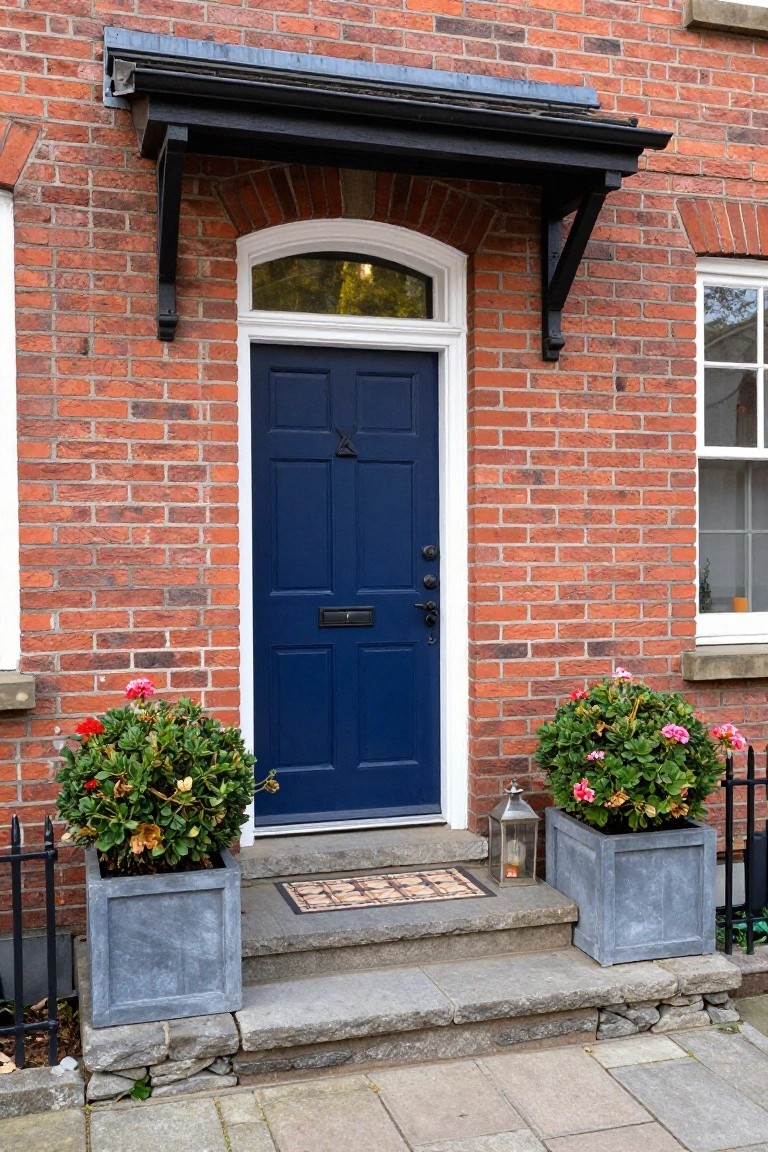 Red brick house exterior with navy blue front door, white arched window above, symmetric gray planters filled with pink geraniums, stone steps, lantern, and tiled doormat.