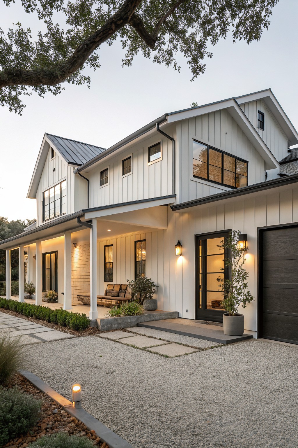 Two-story white board-and-batten sided farmhouse exterior with gray metal roof, wraparound porch, glass entry door, garage door, gravel driveway with pavers, and low plantings at dusk.