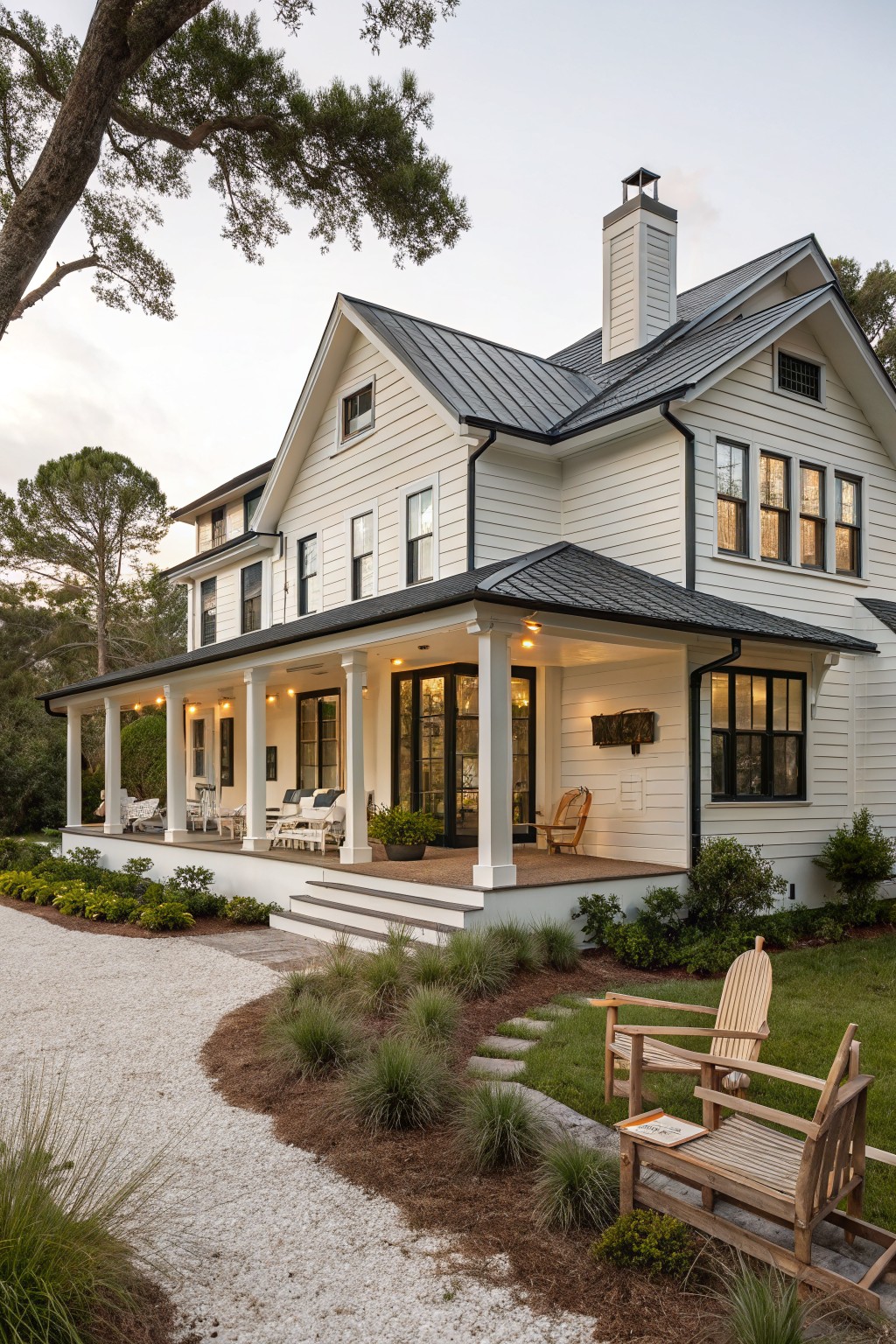White clapboard-sided farmhouse exterior with dark gray standing seam metal roof, wraparound porch with columns and furniture, gravel pathway, and native landscaping under live oak trees.