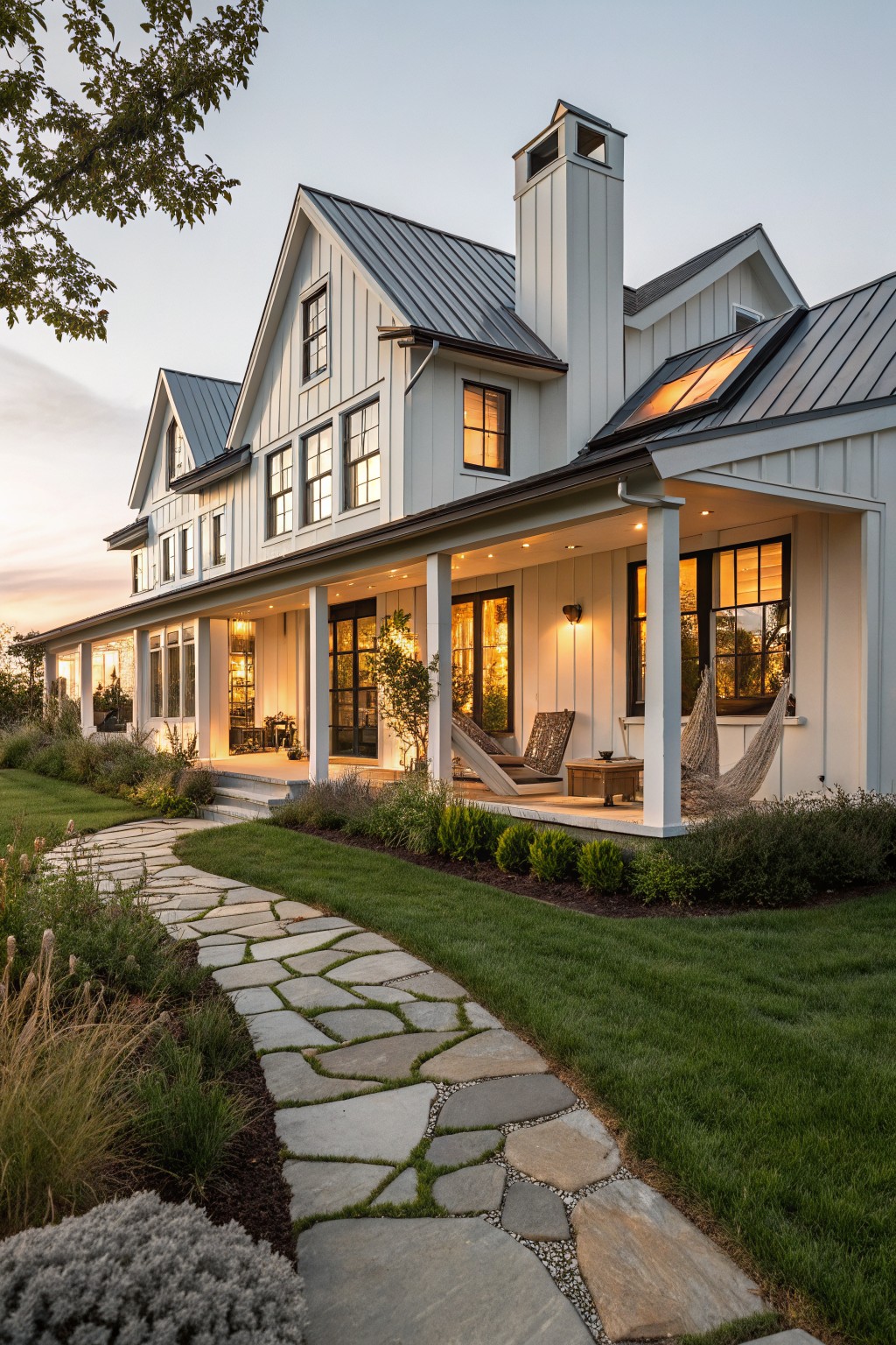 White board-and-batten modern farmhouse exterior with gabled standing-seam metal roof, wraparound porch with hammocks, chairs, and table, stone path through lawn and plantings at dusk.