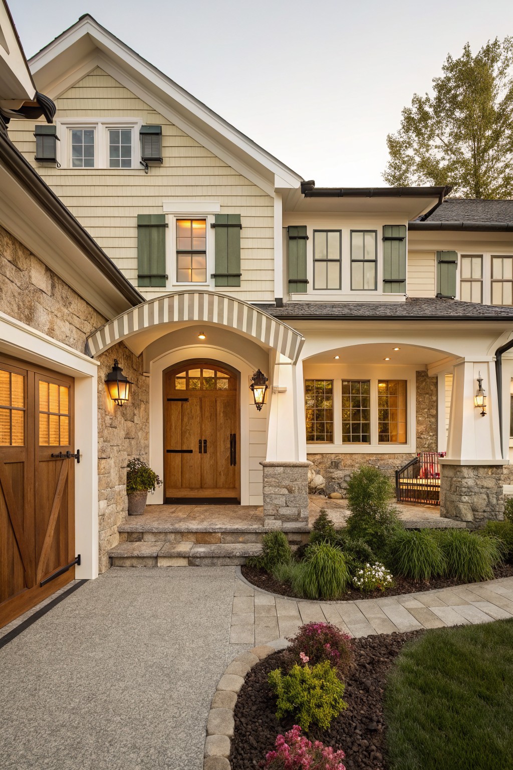 Modern farmhouse house exterior with cream clapboard siding, dark green shutters on multiple windows, light stone foundation and accents, wooden garage door and arched entry door, covered porch with lanterns, and low landscaping along a stone pathway.