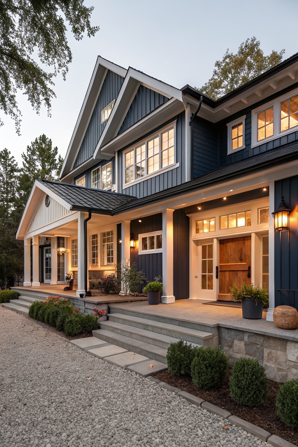 Two-story modern farmhouse exterior with navy blue board-and-batten siding, white trim and porch columns, wooden paneled front door, lit lanterns, steps leading to porch, and gravel driveway edged with shrubs and stone.