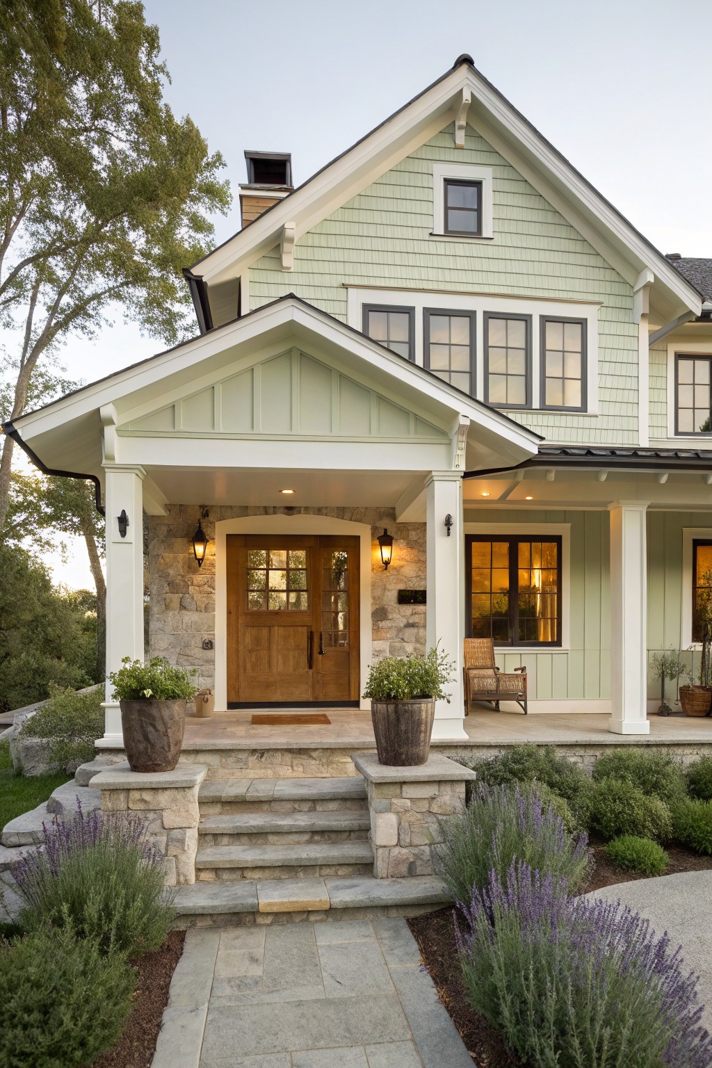 Modern farmhouse exterior with light sage green shingle siding, white gabled porch supported by stone pillars, wooden front door with glass panels, black-framed windows, potted plants on porch, and lavender shrubs along stone steps.