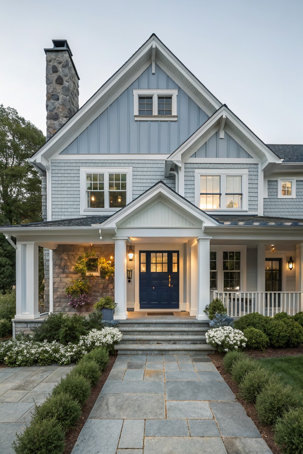 Two-story house with light blue-gray shingle siding, white trim, gabled roof, stone chimney, wraparound porch, navy double front doors, stone steps, bluestone pathway edged with low shrubs and white flowers.