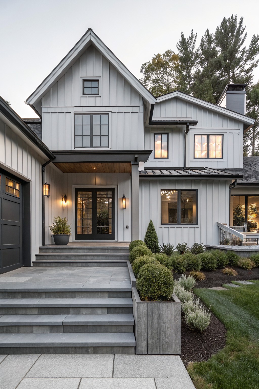 Two-story modern farmhouse house exterior with white board-and-batten siding, black-framed windows and doors, covered entry porch, dark garage door, gray stone steps, and landscaping with shrubs, grasses, and mulch beds.