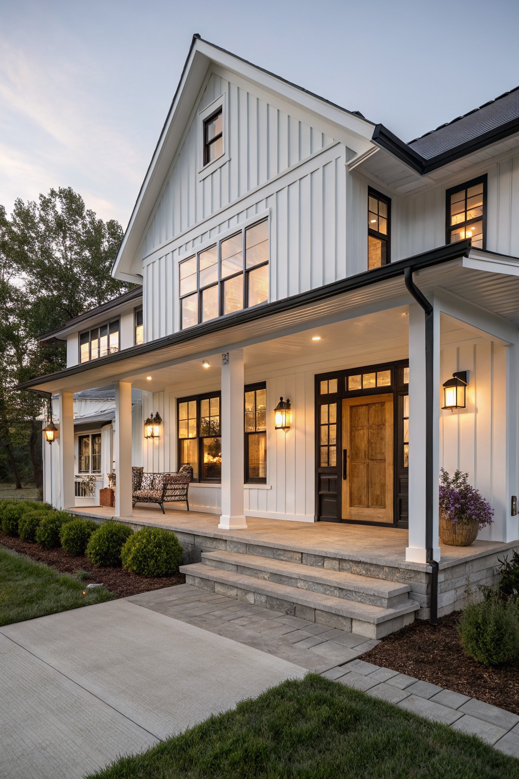 Two-story modern farmhouse with white board-and-batten siding, black window frames and trim, dark shingled roof, covered front porch with wood door and lanterns, steps to entry, and low shrubs in yard at dusk.