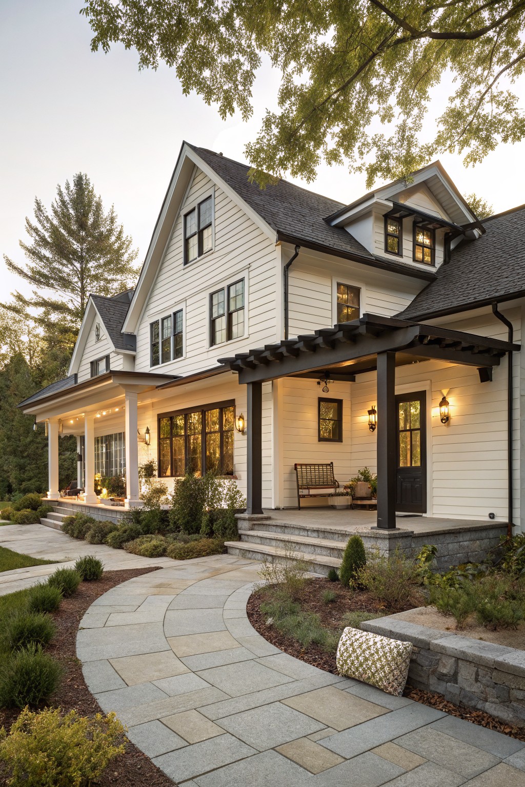 A two-story white board-and-batten house with black window frames, dark shingled roof, pergola-covered entry porch with bench and lanterns, stone steps, and curved bluestone pathway with low plantings.