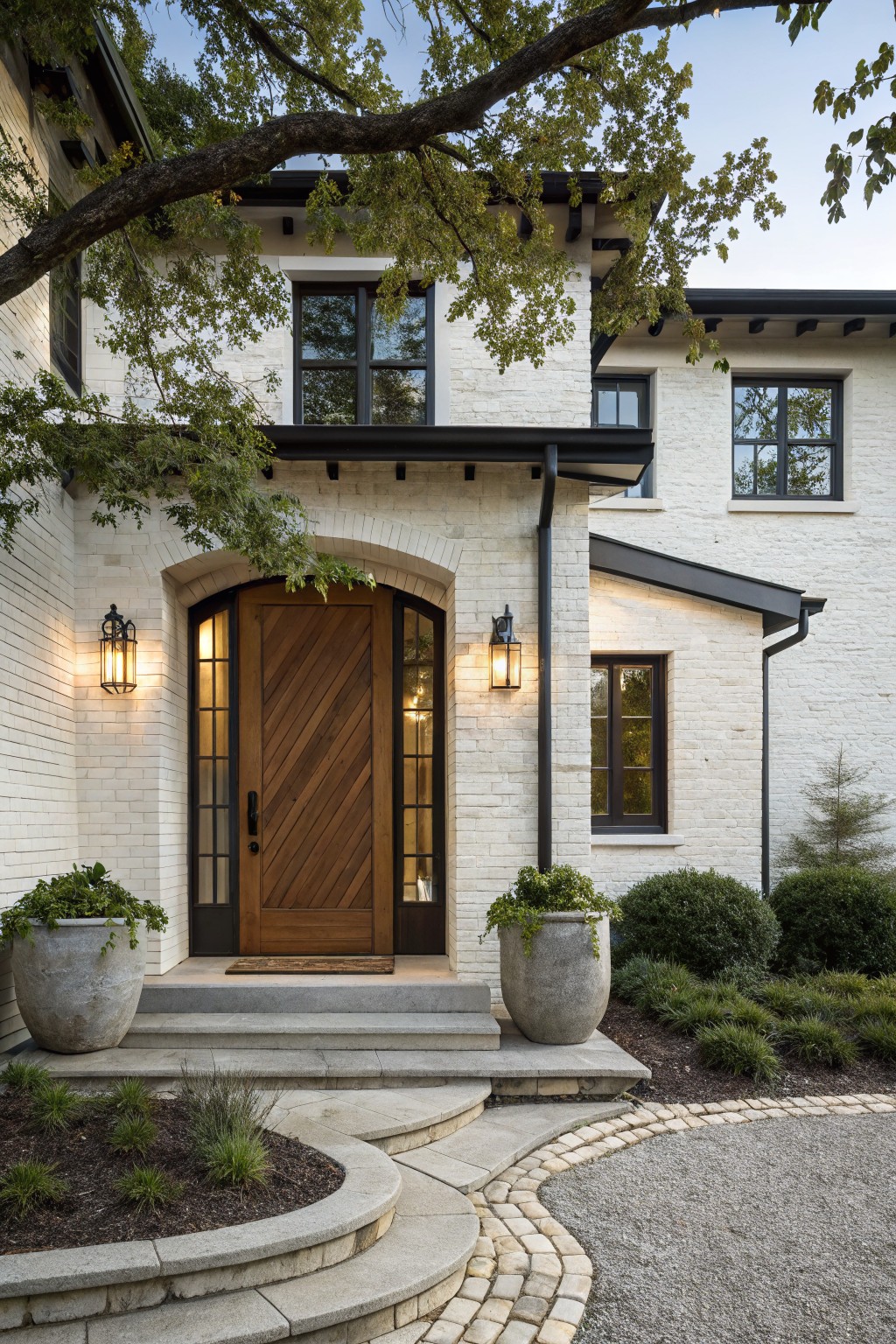White brick house facade with black trim and an arched entry containing a dark chevron-patterned wood door, flanked by lanterns, black-framed windows, stone steps, potted plants, shrubs, and a curved gravel path.