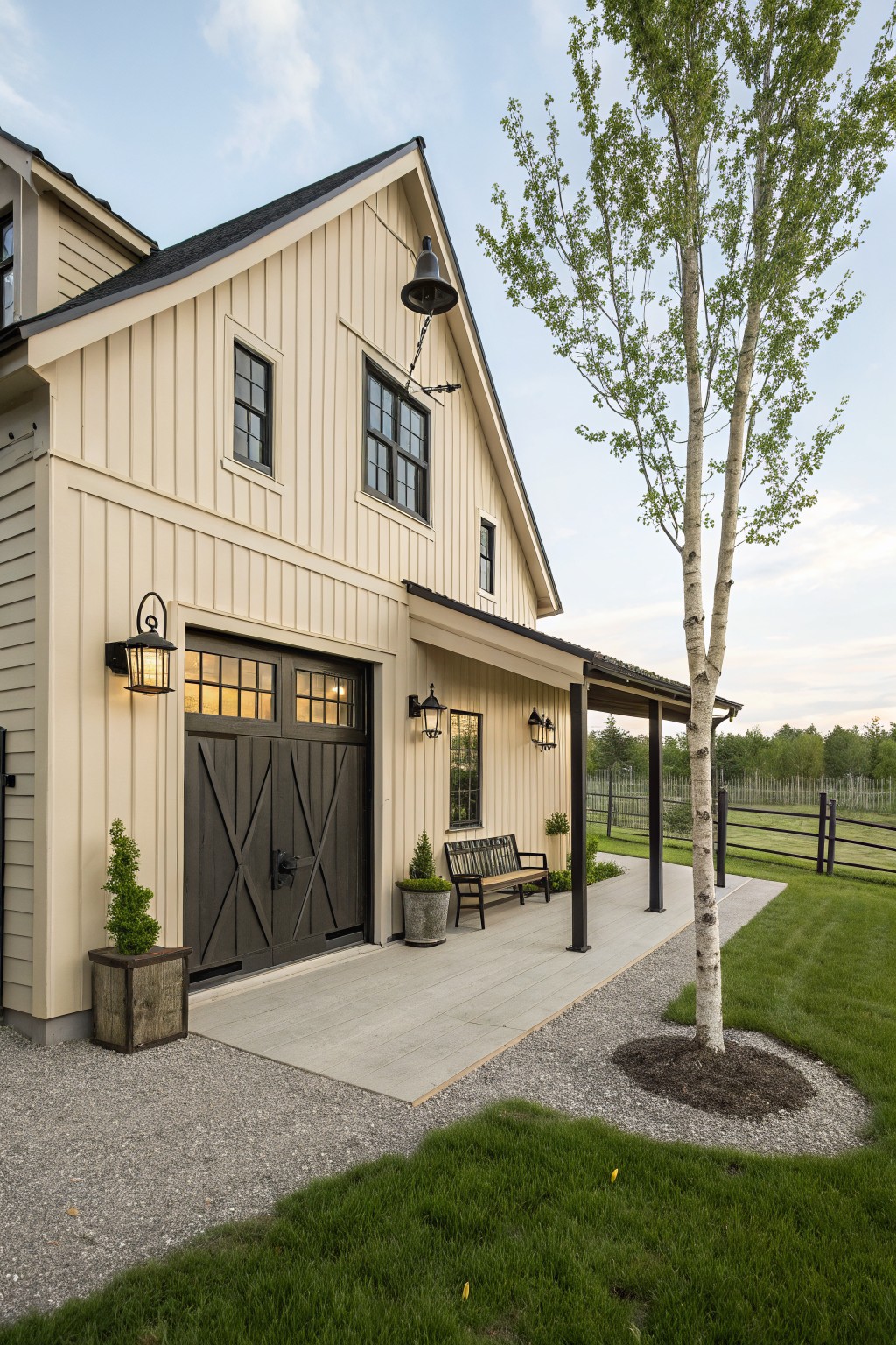 Beige vertical board-and-batten garage building with black X-pattern double garage doors, wall lanterns, covered porch with bench and planters, young birch tree, gravel path, and grass edging a split-rail fence.
