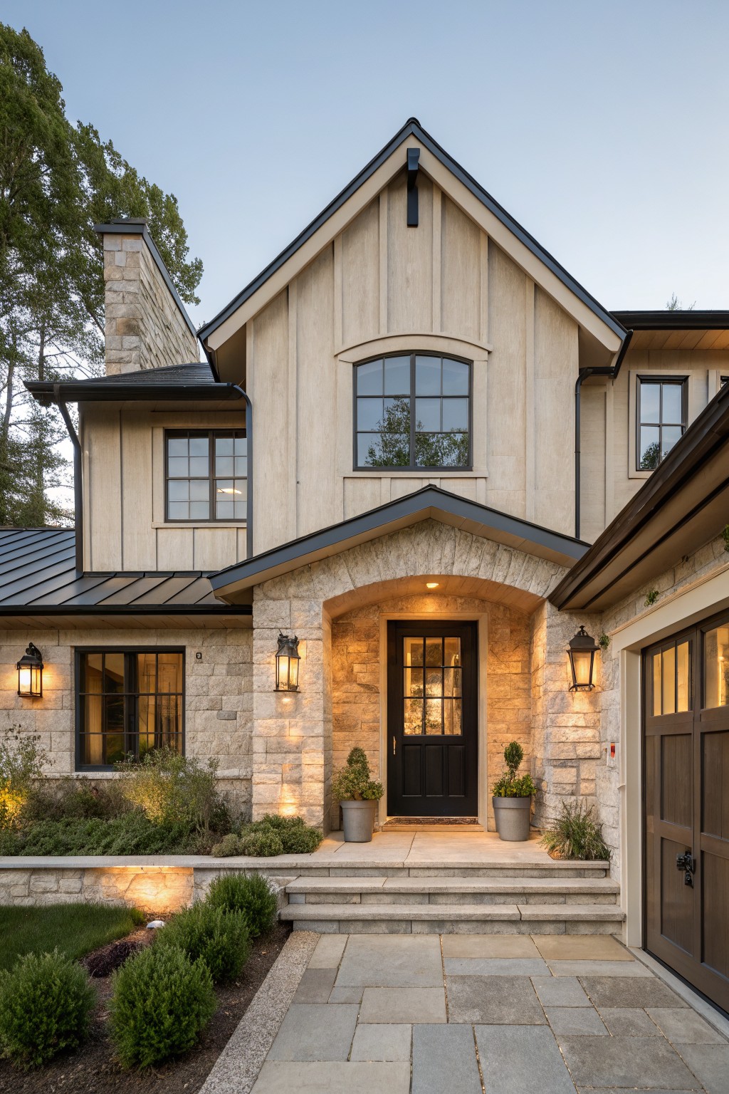 Front exterior of a modern farmhouse home with light wood siding, stone accents including a chimney and arched entryway around a black door, metal roof, lanterns, attached garage door, steps, low shrubs, and bluestone pavers.