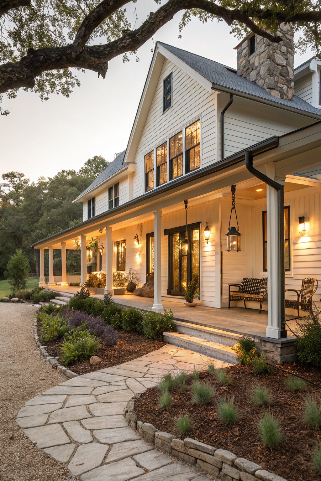 White modern farmhouse with gabled roof, tall stone chimney, wrap-around porch with lanterns and seating, steps leading from gravel pathway edged by plants.