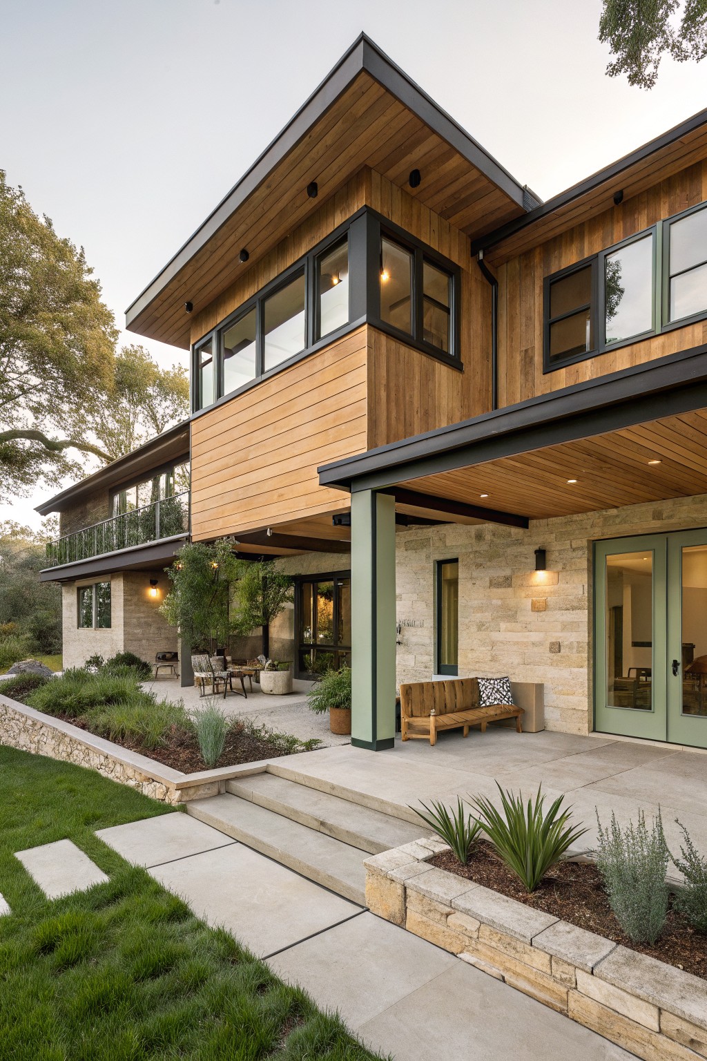 Side exterior view of a two-story modern house with wood-clad upper walls and corners, stone lower walls and foundation, large windows and doors, covered entry patio with bench and plants, and landscaped yard with steps and grass.
